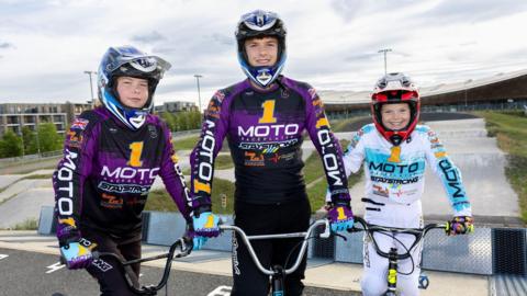 The three siblings stand with their bikes with the BMX track behind them. Katie and Thomas wear purple racing suits with a blue helmet and Phoebe wears a white and blue suit with a red helmet