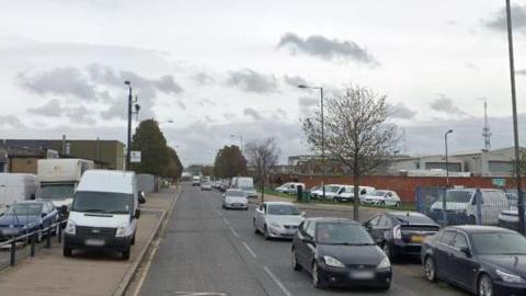A general view of Nathan Way in Thamesmead. There are cars and vans parked on either side of the road. 