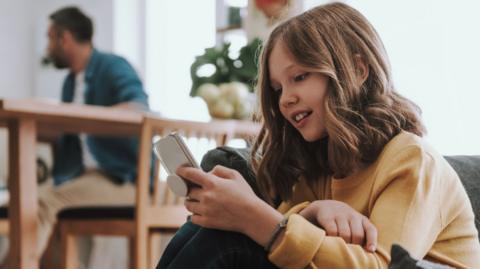Young girl in yellow jumper stares at a mobile phone. her dad can be seen in the background