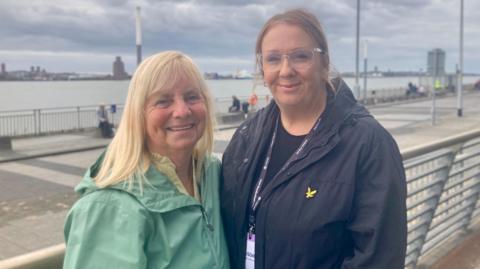 Two women stand by the river Mersey. One is blonde and wears a pale green light anorak. The other women is taller, with long hair tied into a ponytail. She has glasses and is wearing a navy blue jacket. They are both smiling at the camera.