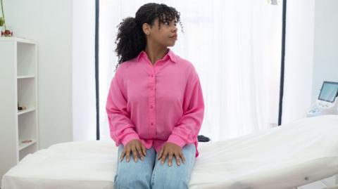 A young woman wearing jeans and a pink shirt sits on a bed waiting for her cervical screening