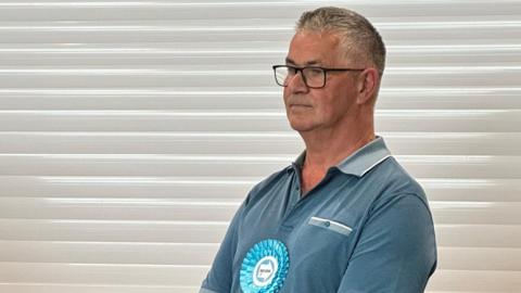 A man stands side on to the camera. He wears a blue T-shirt, with a blue Reform UK rosette. In the background is a white shutter and a beige bar. 