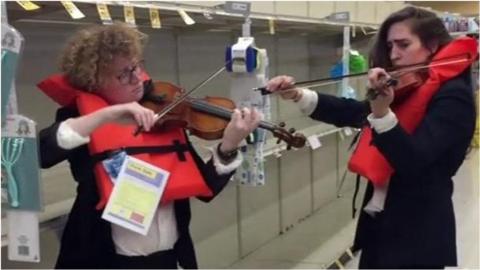 Two women play violins in a supermarket wearing lifejackets