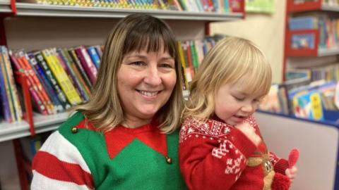 Smiling woman with long fair hair wearing a green, white and red Christmas jumper holds a young girl wearing a red sweater