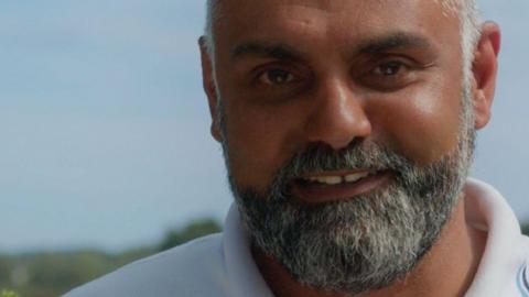 Bal Padda smiles at the camera in a promotional, close-up image. He is standing outside under a light blue sky. He has a grey and white beard and wears a white top.