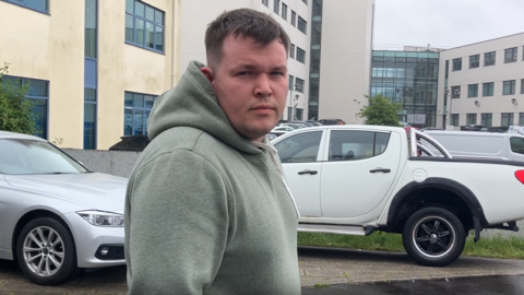 A man wearing a light green hoodie looks at the camera. There are multiple cars parked behind him as well as a cream and white coloured building.
