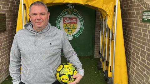 Ashford United co-owner Lloyd Hume holding a yellow football and standing in front of the stadium's tunnel. The tunnel is made up of a yellow fabric canopy and a door with the club's crest on it.