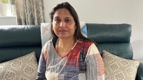 A woman with dark hair and a multiple colour jumper siting on a green sofa inside her living room.