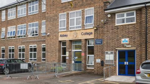 Outside Abbey College. It is a brick building with cars parked outside. Above the blue front entrance door is the school's name and crest.