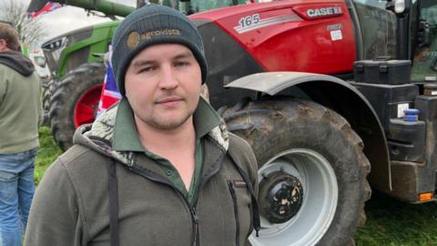 Luke Mason wearing a dark blue beanie and a grey fleece jumper over a green shirt. He is standing in front of a red tractor and looking at the camera with a disappointed facial expression. 