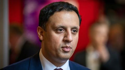 Anas Sarwar, who has dark hair, photographed in a close-up shot. He is visible from the shoulders up, and is wearing a navy suit and white shirt. 