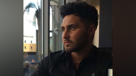 Matthew McIlroy is sitting in front of a large window. He has dark hair which is styled upwards into a quiff and is shorter on the sides. He has a dark beard which is closely trimmed and is wearing a black shirt with the collar opened. 