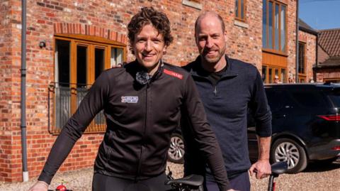Greg James - a man with brown curly hair smiling into the camera and wearing a mic and black zip up jacket and leggings with both of his hands on a tandem in front of him. Next to him is Prince William, smiling and wearing a blue half-zipped up fleece and blue joggers, with one hand on the bike seat and the other on Greg's shoulder. Behind them is the back of The Peppered Pig pub with a brick exterior and long windows. In the corner people can be seen looking on.