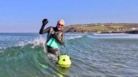 Martina Morgan in the sea holding onto her floatation device and flippers. She is standing in a middle of a wave smiling. She is wearing a wetsuit, a swim cap and goggles. 