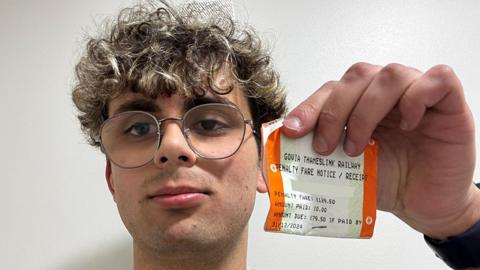 Theo Griffiths with curly dark hair and glasses holds his penalty notice up to the camera