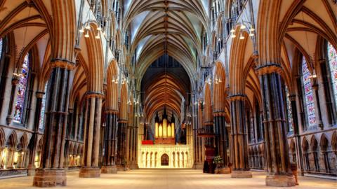 The empty symmetrical nave inside a large, lit-up grand cathedral which has large brown pillars on each side and stained glass windows.