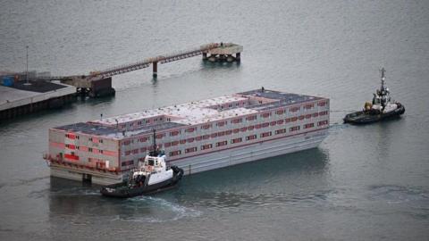 Closer image of the Bibby Stockholm as it leaves Portland. There is a tugboat at the front of it and a second tugboat to the back of it. The Bibby Stockholm is a red and white rectangular barge, moving through the water and there is a dock platform seen to the left of the image.