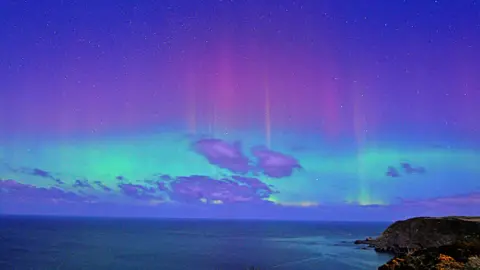 Purple and bright green aurora shimmer in a blue evening sky over the sea and headlands