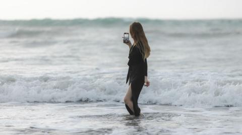 A woman with long dark blonde hair stands in the ocean. She is facing away from the camera and holding her phone in her right hand, she is recording the view of the ocean waves. The woman wears a long sleeved black dress, which has a long side split in the skirt showing her left leg.