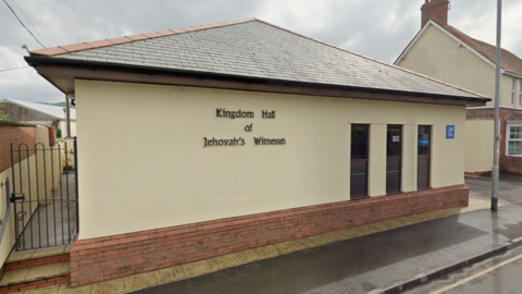 The Kingdom Hall, which is a one-storey building rendered with pale yellow plaster. It has three small windows, a sign which says "Kingdom Hall of Jehovah's Witnesses" and a blue plaque which says JW.org.