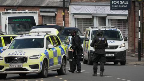 Two armed police officers in bullet-proof vests and wearing black helmets stand next to three police cars and a police van on the street