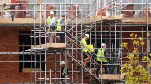 Four men climb down scaffolding stairs on the side of an under-construction brick building. They are wearing yellow high-vis jackets and hard hats.