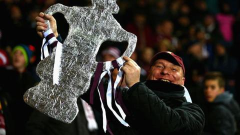 A Northampton Town fan holding up a foil replica of the FA Cup trophy. It has claret and white ribbons attached to it. The fan is wearing a claret baseball cap and has a broad smile