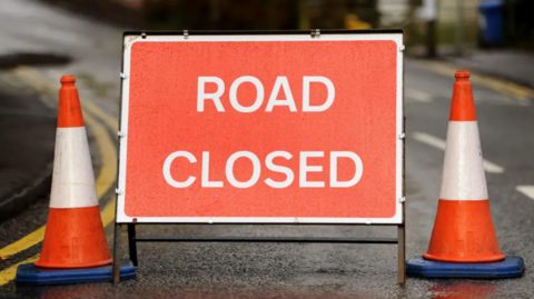 Stock image of a sign reading "road closed". It is red with white capital letters. Orange and white traffic cones are placed either side.