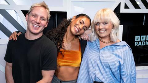 Jamie Laing, Vick Hope and Katie Thistleton in the BBC Radio 1 studio. Jamie is a white man in his 30s with short blonde hair and wears a black T-shirt. Vick is a black woman in her 30s and has her arm around Jamie, her head tilted to the side as she smiles. She has her long hair tired back in a pony tail and wears an orange velvet crop top and black cardigan. Katie is a white woman in her 30s with shoulder length blonde hair. She wears a blue and white striped oversized shirt and smiles at the camera.