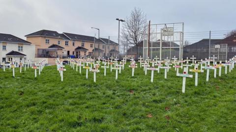 Shows a large number of white crosses in a grassy area with houses in the background, without the large poppy on feature which was stolen