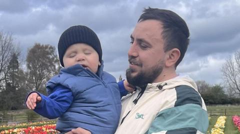 Stepan Kozariichuk is holding baby Bohdan in his arms and looking at him with a smile on his face. They are in a colourful tulip field. Stepan , who has short black hair and a beard, is wearing a white and blue jacket. Bohdan's eyes are closed. he is wearing a blue hat and body warmer.