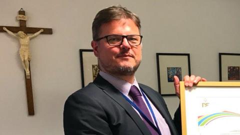 Dean Juric has glasses and a beard, his hair is brown and grey. He is wearing a dark suit with a purple striped shirt and purple tie. In the background is a crucifix depicting Jesus and he is holding a framed Duke of Edinburgh Award certificate.