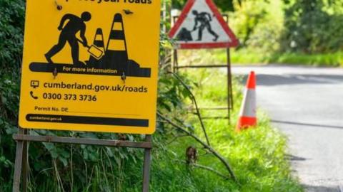 A Cumberland Council yellow road sign which displays a work person digging at the side of the road. There is another highway sign in the distance and an orange traffic cone at the side of the grassy verge.