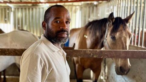 Khaled looks at the camera while standing in front of horse in stable