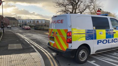 In the foreground, a police van parked in front of a cordon. In the background, a school and blue and white police cordon tape