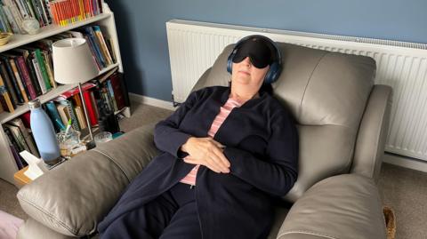 A lady wearing a long dark blue cardigan lies in a grey leather recliner chair in her lounge, wearing a black eyemask and noise-reducing headphones