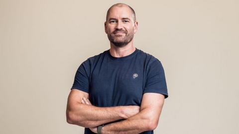 Studio photograph of Paul Saunders with a dark beard and hair wearing a blue T-Shirt with his arms folded