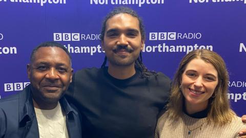 Two men and one woman stand in front of a purple BBC Radio Northampton wall. On the left is Neil who is wearing a navy jacket and white jumper. Leon is in the middle wearing a black t-shirt and sporting a moustache. On the right is Susie who has shoulder length light brown hair and is wearing a cream jumper and a gold necklace with a pendant. All our smiling at the camera.