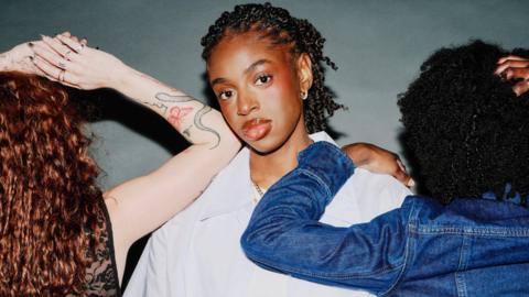 Cat Burns in a promotional shot for her new single. Cat wears an oversized white shirt, gold chain, and has her braided hair tied back. Two women, facing the opposite direction, stand on either side of her. On the left, a red-headed woman with long curly hair rests her tattooed arm on Cat's shoulder while on her right, a woman with curly dark hair wearing a denim jacket rests her hand at the top of her back. 