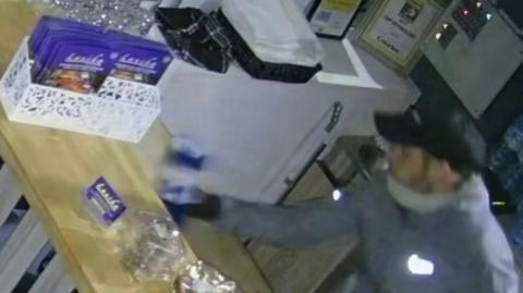 Man in grey jacket, gloves, cap and face covering steals a blue charity box from a restaurant counter in Halifax, January 2026. 