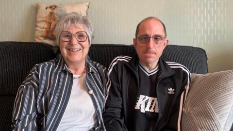 a picture of wayne who's wearing a black tracksuit top, shirt, glasses and short hair.
His mum karen is on the left wearing a striped shirt over a white blouse, she has short-ish white hair and hexagonal glasses - she's smiling a broad smile.

