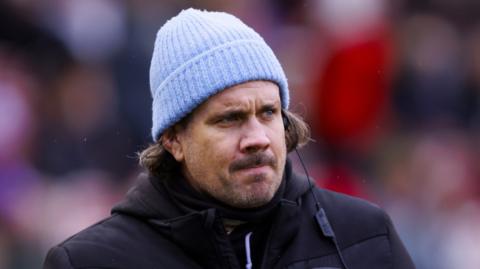 Gloucester-Hartpury head coach Dan Murphy looks on while wearing a black jacket and a blue hat before a Premiership Women's Rugby match.
