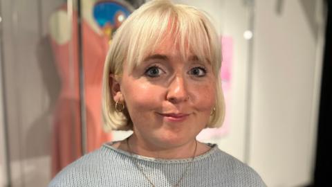 A woman with a white blonde bob looks at the camera. She wears a baby blue jumper with a gold chain hanging from her neck. Behind her, a "coal queen" dress is blurred.