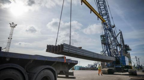 Large steel rods are lifted with a crane. It is a sunny day with blue skies. 