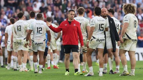 Johann van Graan (centre) holds out his right arm to clap hands with Santiago Carreras, who is walking towards him on the pitch at the end of Bordeaux v Bath. Other Bath players walk around in the background