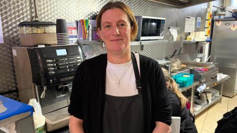 Helen stands in the kitchen, she is wearing a white t-shirt and black cardigan and a black apron over.
