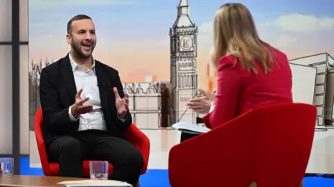 Zack Polanski speaks to Laura K. He is gesturing with his hands in front of him and sits in a red chair. Laura's back is to the camera, she also sits in a red chair and is wearing a red blazer.