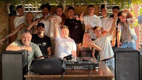 Adam, centre, is surrounded by a group of people behind DJ decks and large speakers. Everyone is smiling with their hands in the air. 