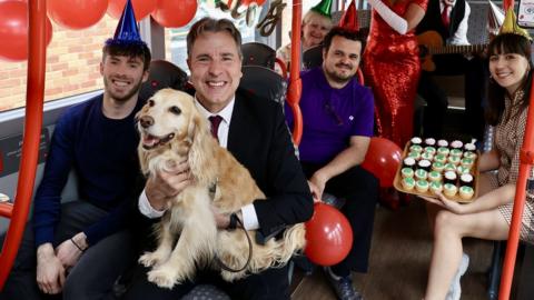 Dan Norris holding a dog on a bus decorated with birthday balloons and bunting