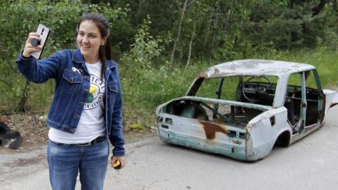 A visitor posses for a selfie next to a wrecked car at the Chernobyl exclusion zone